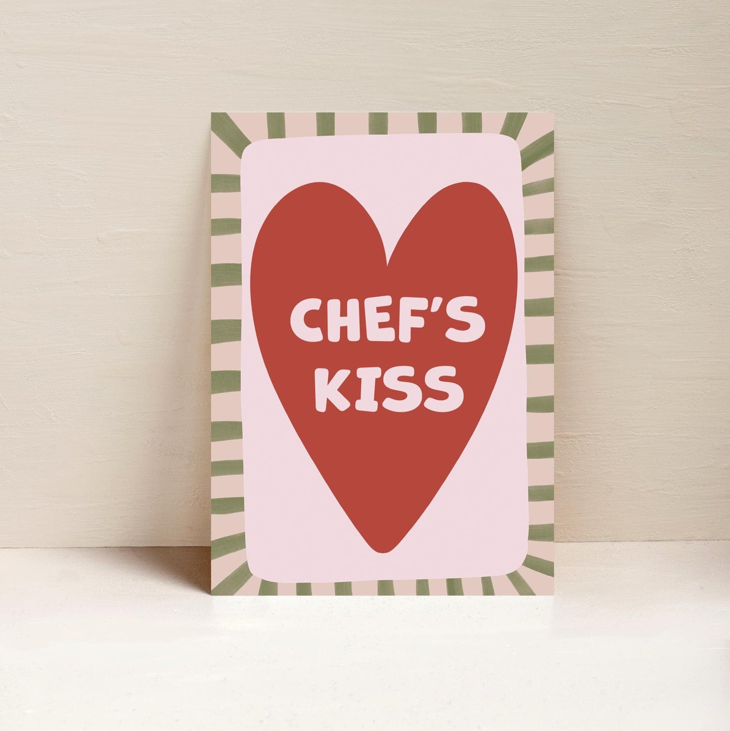 A framed art print with a pink background featuring a red heart and the text 'CHEF'S KISS' in bold white letters, presented against a neutral backdrop.