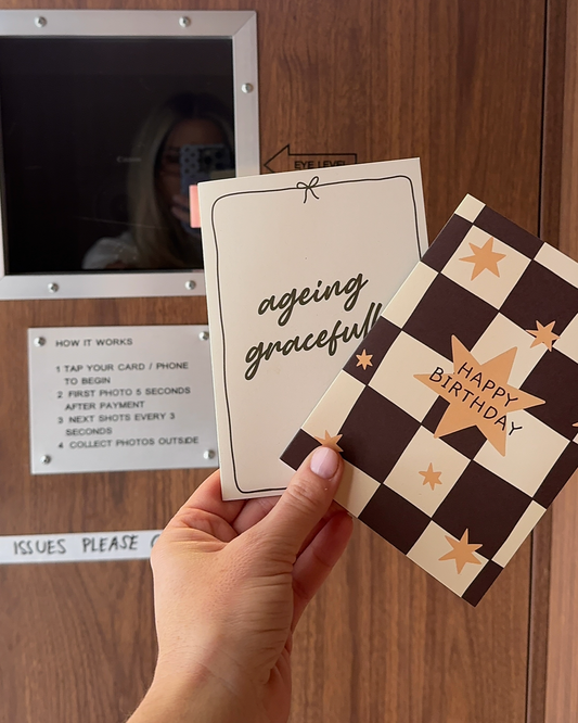 A hand holds two greeting cards in front of a wooden door. One card is cream with black script reading 'aging graceful' and the other has a brown and cream checkerboard pattern with gold stars and 'HAPPY BIRTHDAY' text.